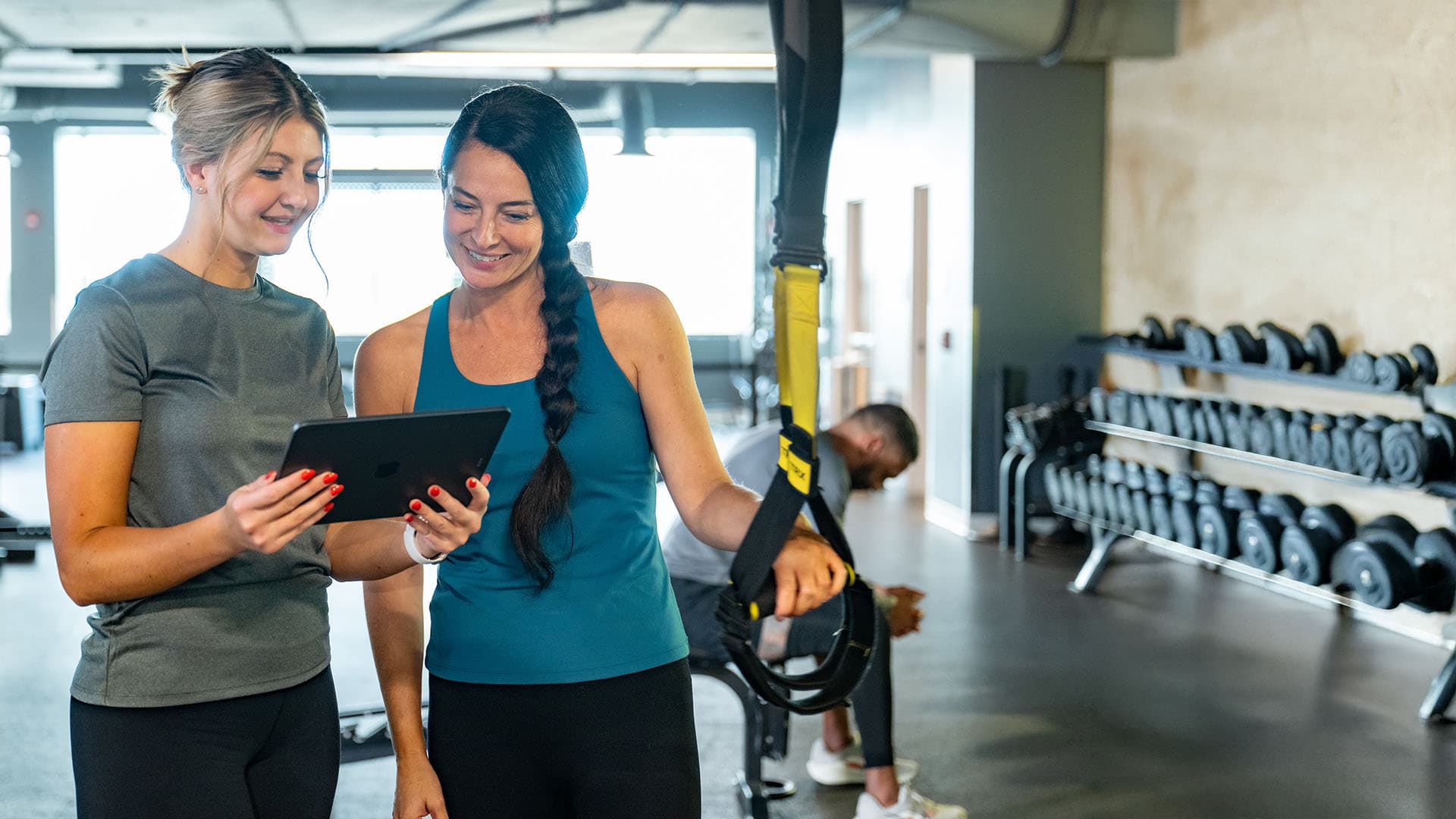 A fitness coach and a woman smile while looking at a tablet together in a gym, with suspension straps and rows of dumbbells in the background.